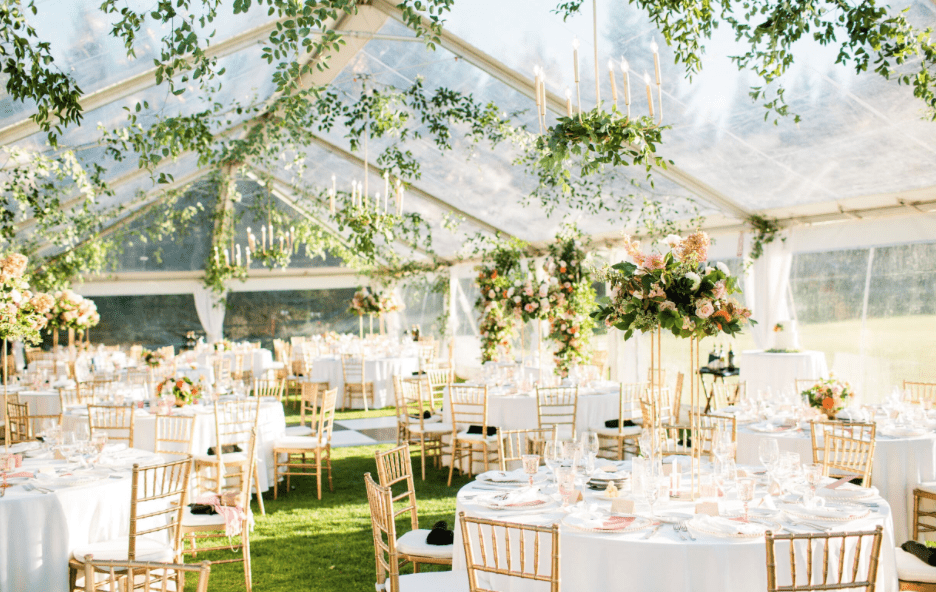 Gold Chiavari chairs arranged around elegant reception tables beneath a clear-top wedding tent with floral centerpieces and greenery.