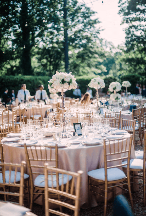 Gold Chiavari chairs surrounding an elegant outdoor wedding reception table with blush linens, white floral centerpieces, glassware, and string lights.