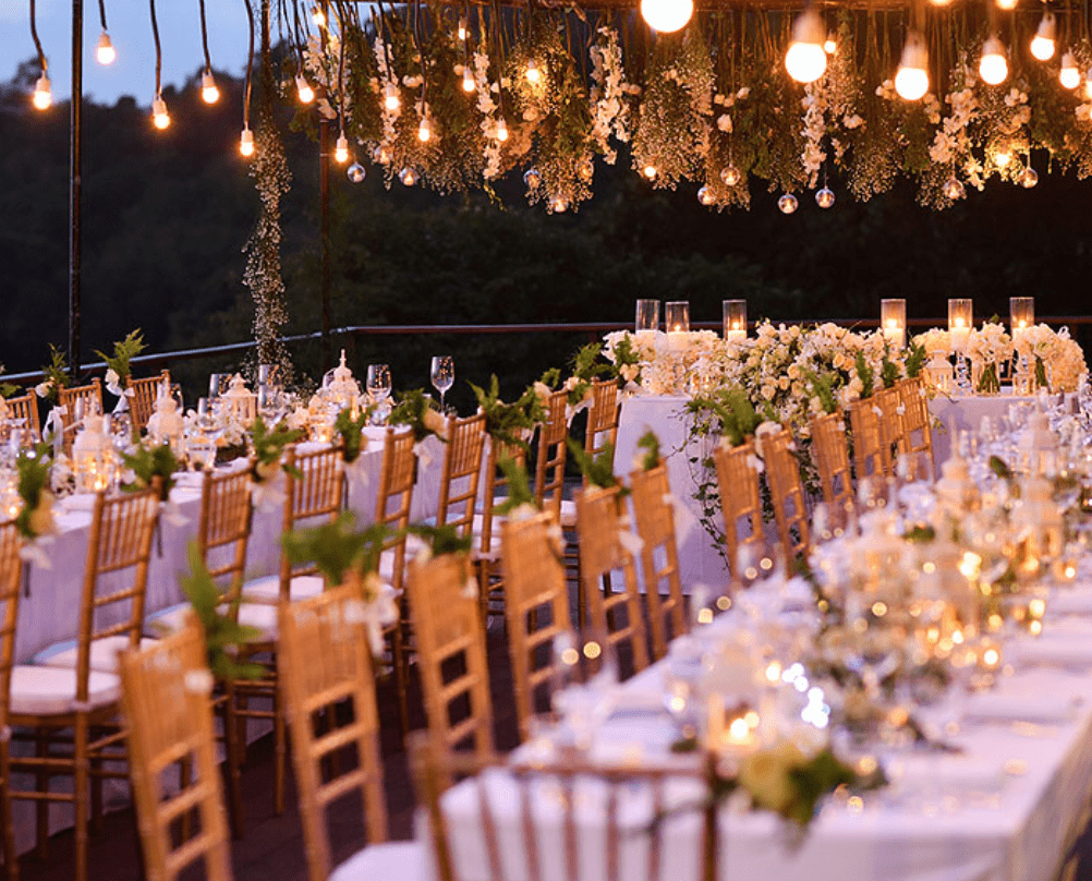 Gold Chiavari chairs surround elegant reception tables beneath hanging lights and floral greenery at an outdoor evening wedding. A warm example of upscale chair rentals for weddings and special events in Middlesex, NJ.