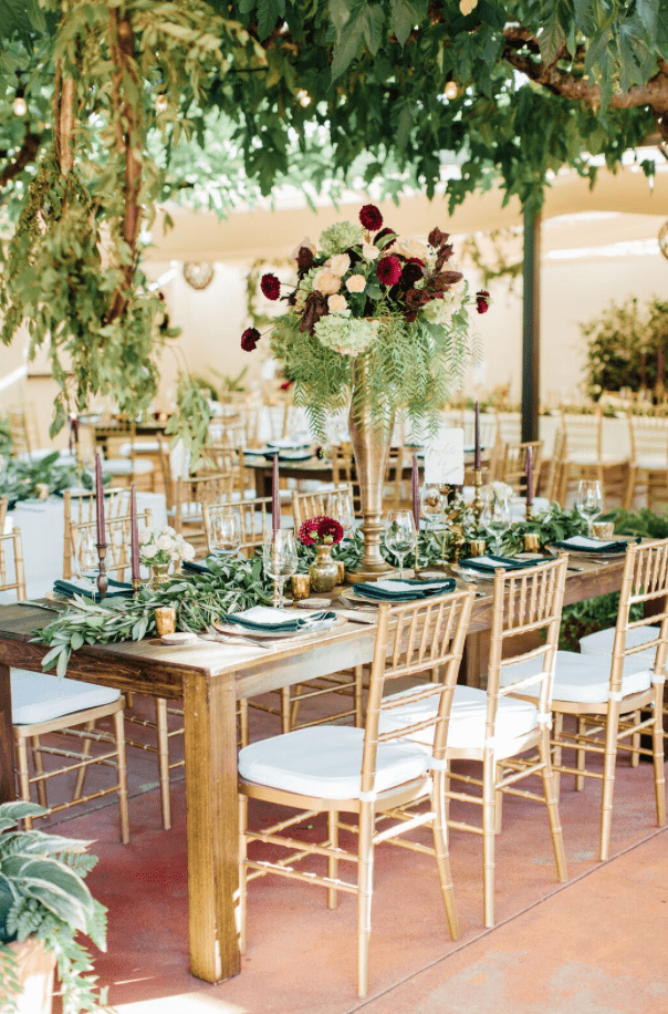 Gold Chiavari chairs paired with rustic wood tables, lush greenery, and burgundy floral centerpieces at an elegant garden-style reception. A beautiful example of mixing classic chair rentals with natural wedding décor.